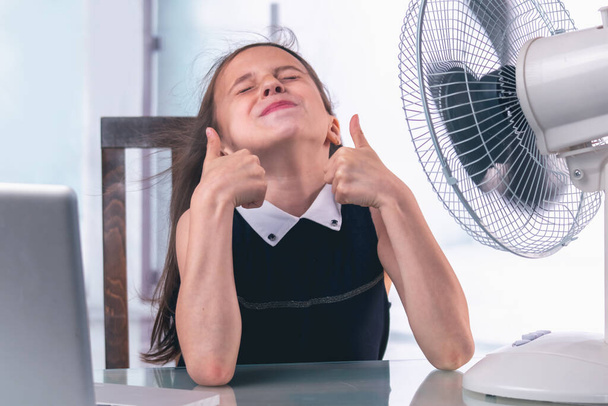 Menina de negócios jovem atraente sentado na mesa no escritório durante uma onda de calor de verão, ela está muito feliz em se refrescar com um ventilador elétrico - Foto, Imagem