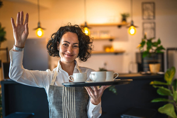 Een volwassen vrouw Kaukasische serveerster in cafe of restaurant dragen lade met koffie vrouwelijke ondernemer op het werk echte mensen kopiëren ruimte klein bedrijf concept - Foto, afbeelding