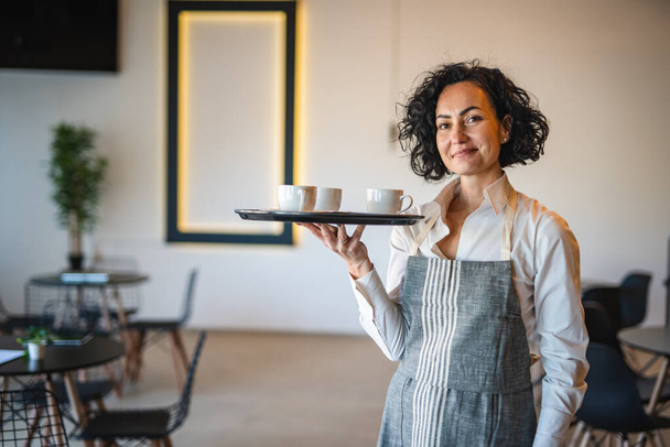 Egy érett nő kaukázusi pincérnő a kávézóban vagy az étteremben tálcát visz a kávéval női vállalkozó a munkahelyen valós emberek másolják tér kis üzleti koncepció - Fotó, kép