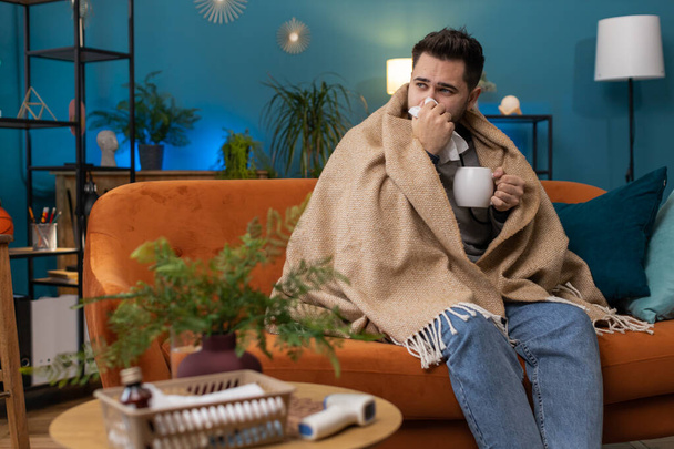Sick man wrapped in plaid sit alone shivering from cold on sofa drinking hot tea in unheated apartment without heating due debt. Unhealthy ill guy feeling discomfort try to warming up at home room - Photo, Image