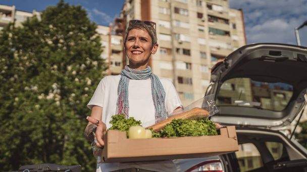 Jedna kobieta dojrzała kaukaska kobieta stoi na parkingu bu samochód przed sklepem spożywczym supermarket trzymając pudełko z jedzeniem i warzywami w słoneczny dzień szczęśliwy uśmiech zdrowe jedzenie wegańskie koncepcja - Zdjęcie, obraz