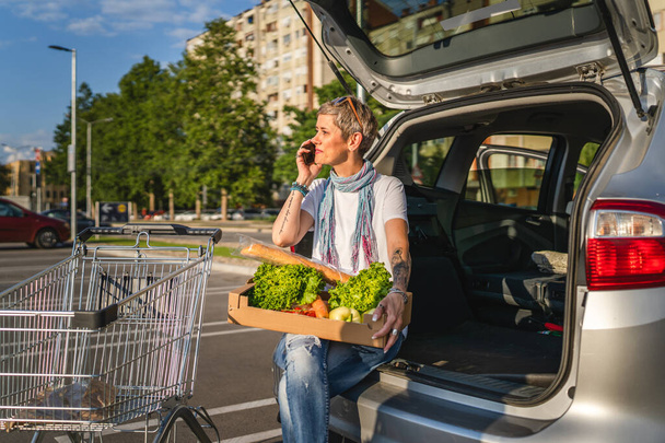Jedna kobieta dojrzała kaukaska kobieta siedzi w bagażniku swojego samochodu na parkingu supermarketu lub sklepu spożywczego z warzywami w pudełku umieszczając je w pojeździe - Zdjęcie, obraz