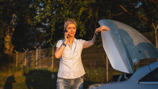 one woman mature female standing on the road in the evening sunset by the broken vehicle car automobile failed engine open hood making a phone call for help roadside assistance towing service concept - Zdjęcie, obraz