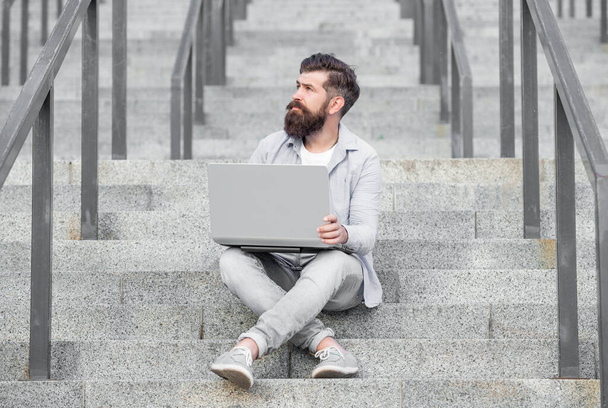 dreamy guy has online video communication sitting on stairs. guy has online video communication on laptop. guy has online video communication outdoor. guy has online video communication outside. - Photo, image