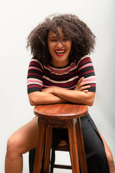 Portrait of a beautiful smiling cheerful young woman with curly hair sitting on a wooden stool. Isolated on light gray background. - Фото, изображение
