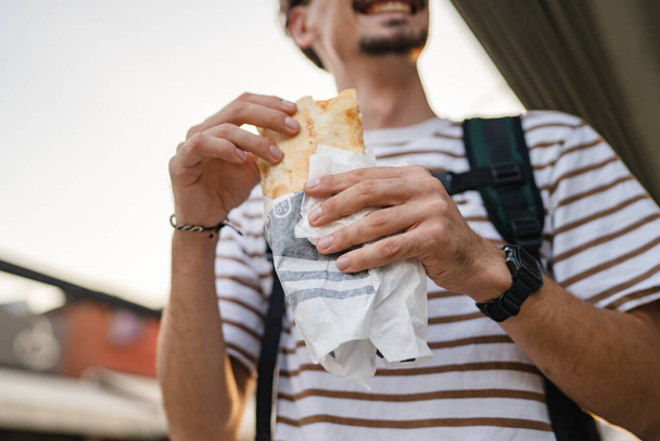 One man young adult modern caucasian male in the city in sunny day walk and eat sandwich fast food concept urban life copy space tourist eating on the street real person - Fotoğraf, Görsel