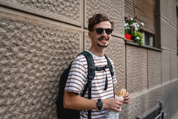 Один чоловік молодий дорослий сучасний кавказький чоловік у місті в сонячний день стоїть і їсть сендвіч концепцію швидкого харчування міське життя копіювання простору турист їсть перед будівельною стіною реальний портрет людини
 - Фото, зображення