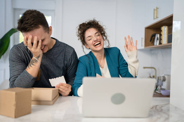Young caucasian couple woman and man wife and husband or girlfriend and boyfriend opening gifts box presents and read card in front of laptop computer at home having online video call happy copy space - 写真・画像