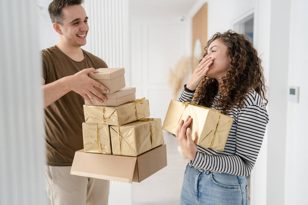 γυναίκα νεαρός ενήλικας καυκάσιος γυναίκα στέκεται στο σπίτι με κουτί δώρου παρουσιάζει κρατώντας στα χέρια, ενώ ο φίλος ή ο σύζυγός της ο άνθρωπος δώσει κουτιά ευτυχισμένο χαμόγελο πραγματικοί άνθρωποι γιορτάζουν έκπληξη έννοια - Φωτογραφία, εικόνα