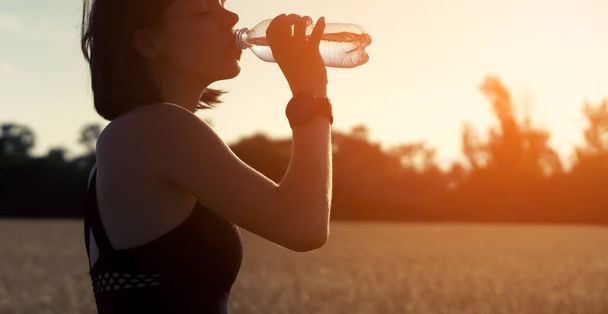 Молода дівчина робить перерву під час інтенсивних тренувань і бігу і п'є освіжаючу прохолодну воду з пляшки, жінка тренується надворі ввечері на заході сонця
. - Фото, зображення
