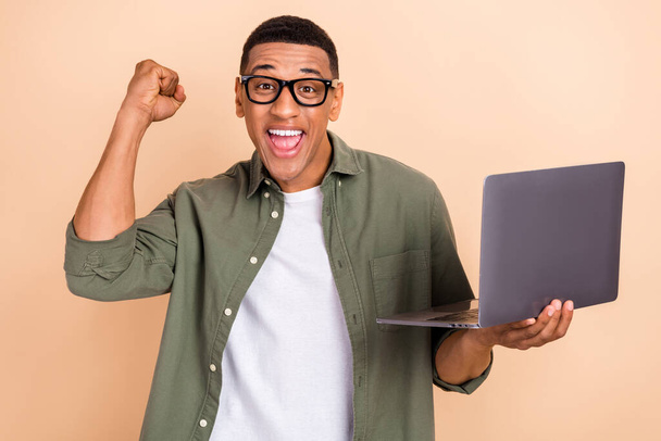 Photo of delighted positive guy hold use netbook raise fist achievement accomplishment isolated on beige color background. - 写真・画像