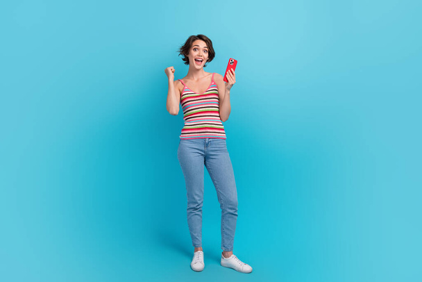 Full body portrait of delighted crazy lady hold smart phone raise fist success achieve isolated on blue color background. - Valokuva, kuva