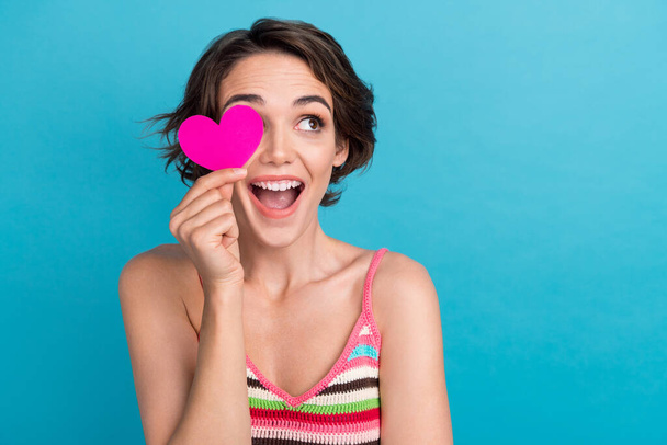 Photo of funny young girl bob hair wear singlet surprised look cover her eye pink postcard heart symbol isolated on blue color background. - 写真・画像