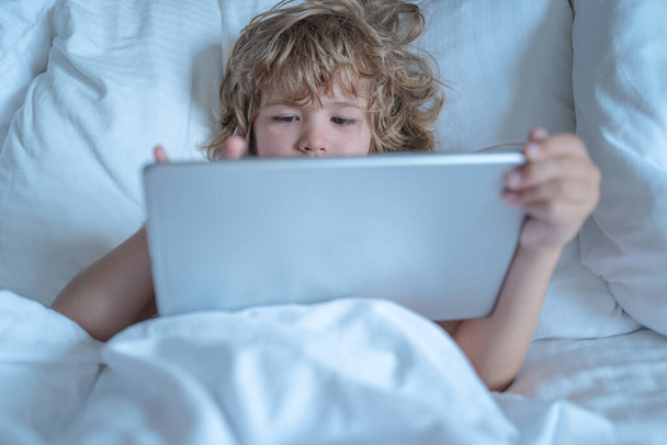 Child in bed under a blanket plays on a tablet in morning. The childs face by a tablet. Child using tablet in bed at morning. Kid in morning bedroom watching movie on tablet - Foto, Imagen