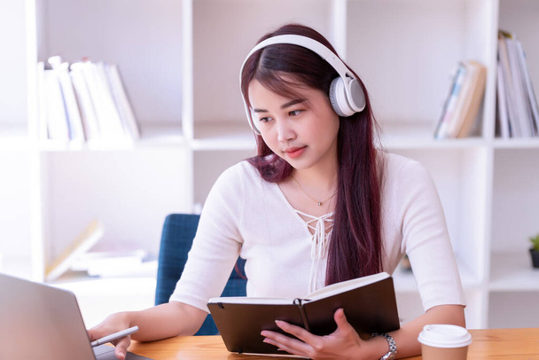 Asian women wear headphones listen to music relax and write notes. Pretty woman learning online. She puts on headphones and works. - Zdjęcie, obraz