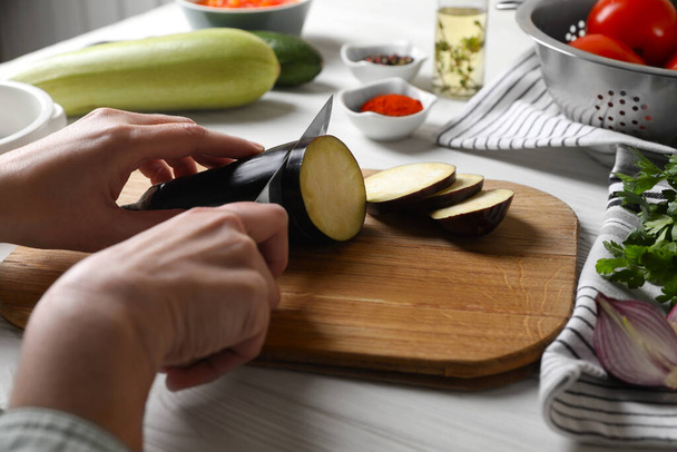 Cooking delicious ratatouille. Woman cutting fresh eggplant at white wooden table, closeup - Foto, afbeelding