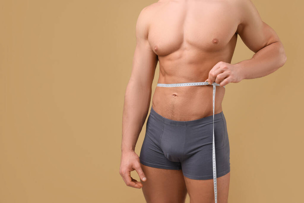 Athletic man measuring waist with tape on brown background, closeup and space for text. Weight loss concept - 写真・画像