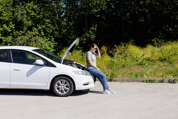 ブルネットの女性は助けを待っている、オープンフード付きの白い壊れた車の上に座っている - 写真・画像