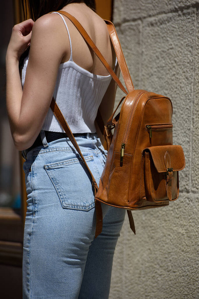 beautiful brunette girl in a blue jeanse and white T-shirt posing with a light brown leather backpack. street photo - Фото, изображение