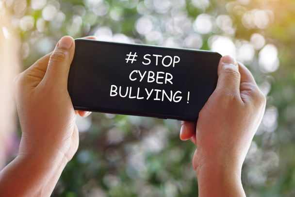 Zamknij ręce trzyma smartfona ze słowem # Stop cyber nękanie na ekranie. Concept, wzywanie wszystkich do zaprzestania znęcania się za pomocą brzydkich słów i złych sms-ów w mediach społecznościowych online lub na platformach wiadomości.   - Zdjęcie, obraz