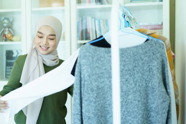Happy cheerful Asian young muslim woman choosing a clothes in fitting room on a clothing hanger. Modern lifestyles concept of muslim people. - 写真・画像