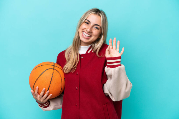 Joven mujer jugadora de baloncesto caucásica aislada sobre fondo azul saludando con la mano con expresión feliz - Foto, imagen