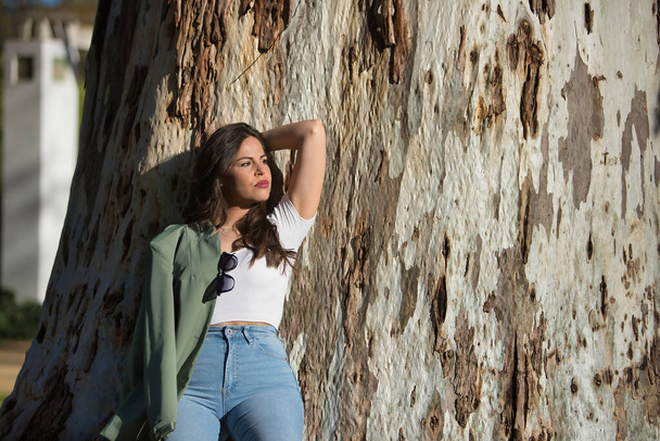 Young and beautiful woman, Hispanic, brown hair, with green jacket and jeans, looking at infinity, relaxed and calm, leaning on the trunk of a huge tree. Concept of solitude, relax, calm. - Photo, image