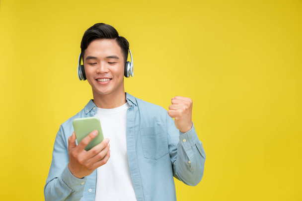 Delighted jonge Aziatische man het dragen van technologie draadloze hoofdtelefoon voor het luisteren naar muziek via mobiele telefoon - Foto, afbeelding