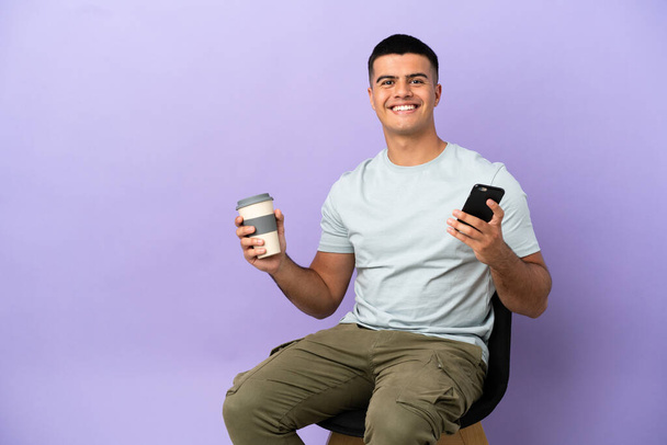 Young man sitting on a chair over isolated background holding coffee to take away and a mobile - Photo, Image