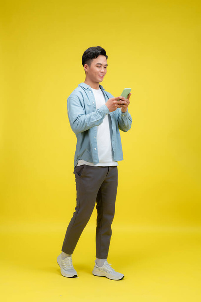Happy man in casual kleding met behulp van mobiele telefoon chatten online, lopen op gele achtergrond - Foto, afbeelding