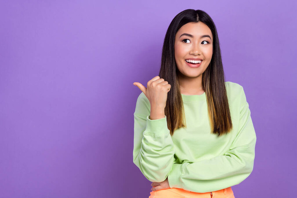Photo of sweet excited girl dressed green sweatshirt pointing thumb looking empty space isolated violet color background. - Photo, Image