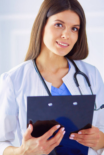 Médecin femme debout avec un dossier dans les mains à l'hôpital, souriant à la caméra - Photo, image