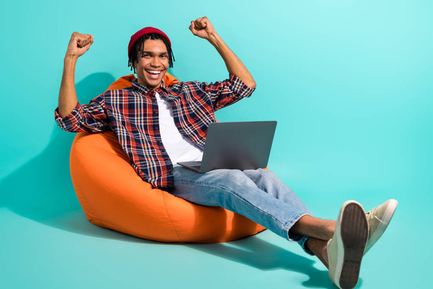 Full length photo of ecstatic guy wear hat sit on bean bag with laptop raising fists up win bet isolated on turquoise color background. - Fotó, kép