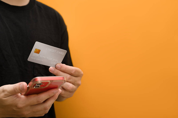 Sarı arka planda kredi kartı ve akıllı telefon tutan bir adam var. E-ticaret, bankacılık hizmeti, online alışveriş. - Fotoğraf, Görsel