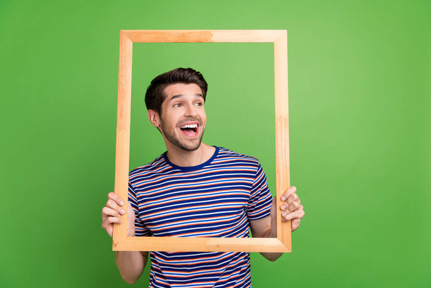 Photo of funny positive young brunet hair guy holding wooden portrait frame capture laughing cadre joke isolated on green color background. - Foto, Imagen