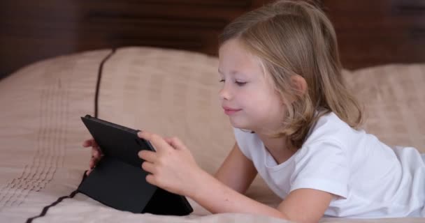 A small girl lying on the sofa looks at the screen of the tablet, close-up. Home schooling, kids cyber security - Footage, Video