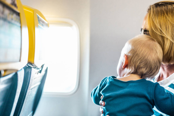 Mamá y su hijo volando en avión. Madre sosteniendo y jugando con su bebé bebé en su regazo durante el vuelo comercial de la economía. Concepto de foto de viaje en avión con bebé. Gente real - Foto, imagen