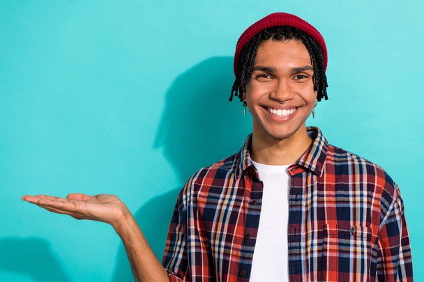 Photo of satisfied cool guy with cornrows dressed checkered shirt arm demonstrate product empty space isolated on teal color background. - Foto, Imagem