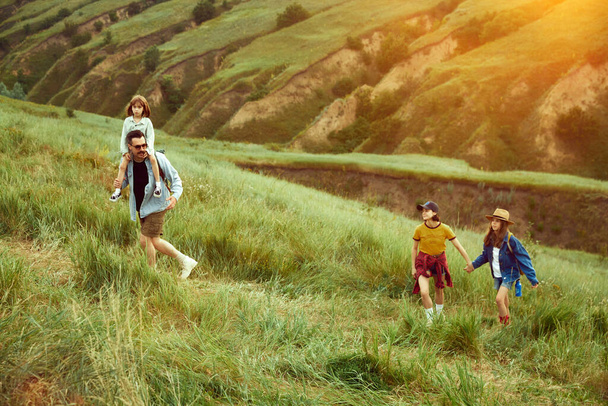 Parents going hiking with their children, daughters on warm summer evening. Beautiful hills landscape. Sunset. Concept of leisure time, fun, nature, active lifestyle, family, travel - Foto, Imagen