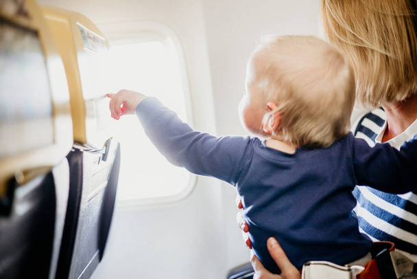 Мама и ребенок летят на самолёте. Мать держит и играет со своим ребенком младенца мальчика на коленях во время экономического коммерческого полета. Концептуальное фото авиаперелетов с малышом. Реальные люди - Фото, изображение