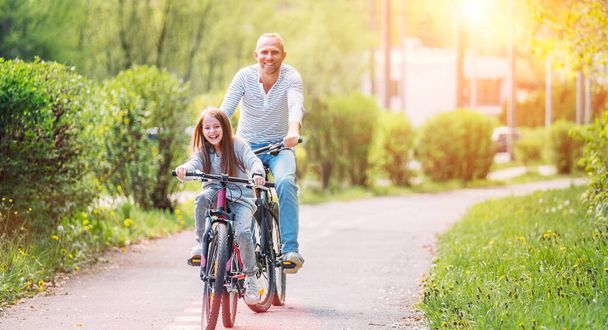 Portraits Smiling father with daughter during summer outdoor bicycle riding. They enjoy togetherness in the summer city park. Happy parenthood and childhood or active sport life concept image. - Foto, Imagen