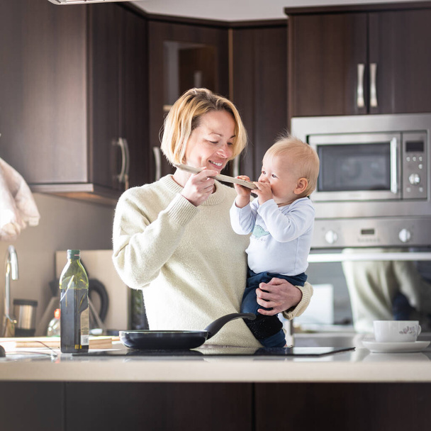 Happy mother and little infant baby boy cooking and tasting healthy dinner in domestic kitchen. Family, lifestyle, domestic life, food, healthy eating and people concept - Fotografie, Obrázek