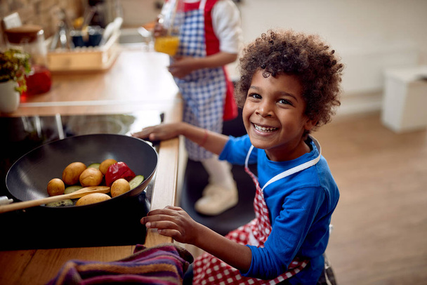 Afro-American boy with a smile on his face, he gazes directly into the camera, exuding a sense of confidence.  In front of him sits a large frying pan filled with vibrant and colorful vegetables. - Фото, изображение