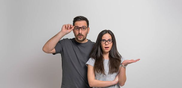 Portrait of worried and scared young couple dressed in casuals looking at camera shockingly. Amazed woman gesturing and man holding eyeglasses while standing against background - Photo, Image