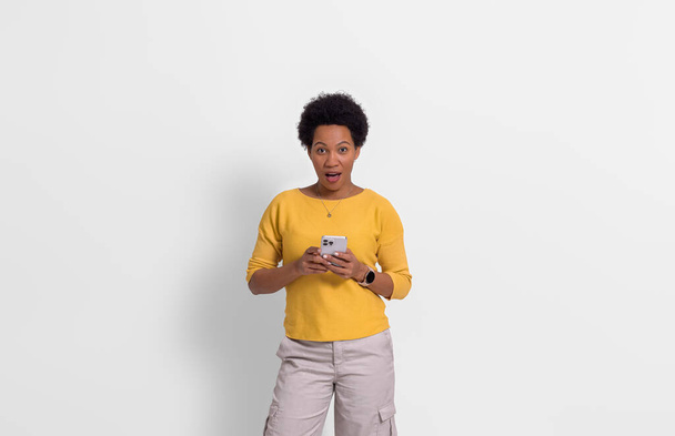 Fiatal nő afro frizura döbbenetesen olvasás üzenetek intelligens telefon fehér háttér - Fotó, kép