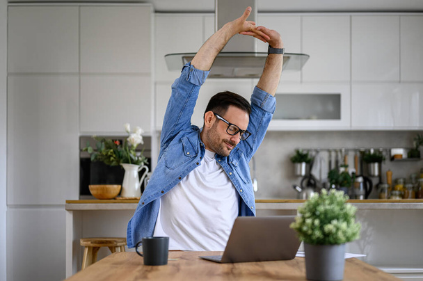 Empresario masculino cansado estirando los brazos mientras trabaja horas extras en el ordenador portátil en el escritorio en la oficina en casa - Foto, imagen
