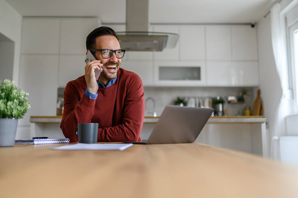 Allegro giovane uomo d'affari che parla su smart phone mentre lavora su laptop sulla scrivania in home office - Foto, immagini