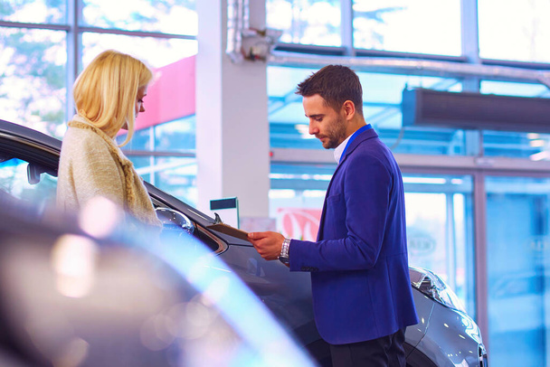 Dealer with woman stands near a new car in the showroom. - Foto, imagen