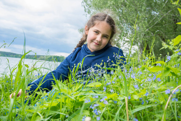 In the summer afternoon, on the river bank, a girl with a pigtail sits in the tall grass. - Photo, image