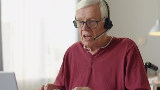 Sklopit záběr staršího muže s sluchátky sedí na invalidním vozíku doma a mluví prostřednictvím videohovoru na notebooku - Záběry, video